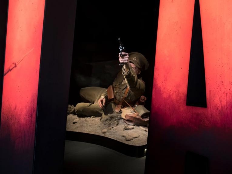 Gallipoli: The Scale Of Our War, 2019. Photo by Maarten Holl. Te Papa (138391) A mmodel of a man in an army uniform lying on the ground of a fake desert pointing a gun in the air.