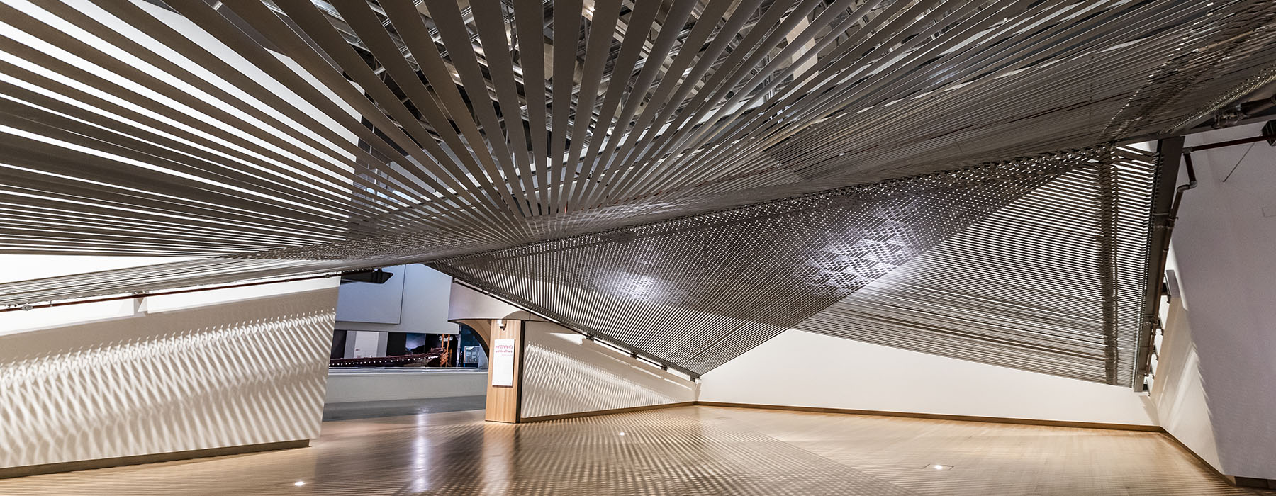 A large-scale, woven installation that is suspended from above and spans a double-height gallery space with a wooden floor and white walls. Woven, grey and reflective tie-downs create shadows and reflections on the walls and floor.