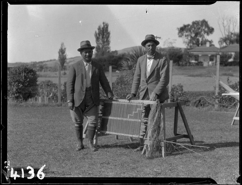 Black and white photo of two people standing in a field next to a piece of weaving