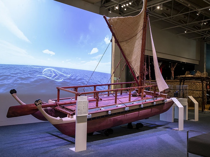 A view of the Manu Rere Moana exhibition within Mana Whenua, 2022. Photo by Maarten Holl. Te Papa (219668) General view of an exhibition space with a large projection on the wall, a waka (Māori canoe) in the foreground, and further exhibits in the background