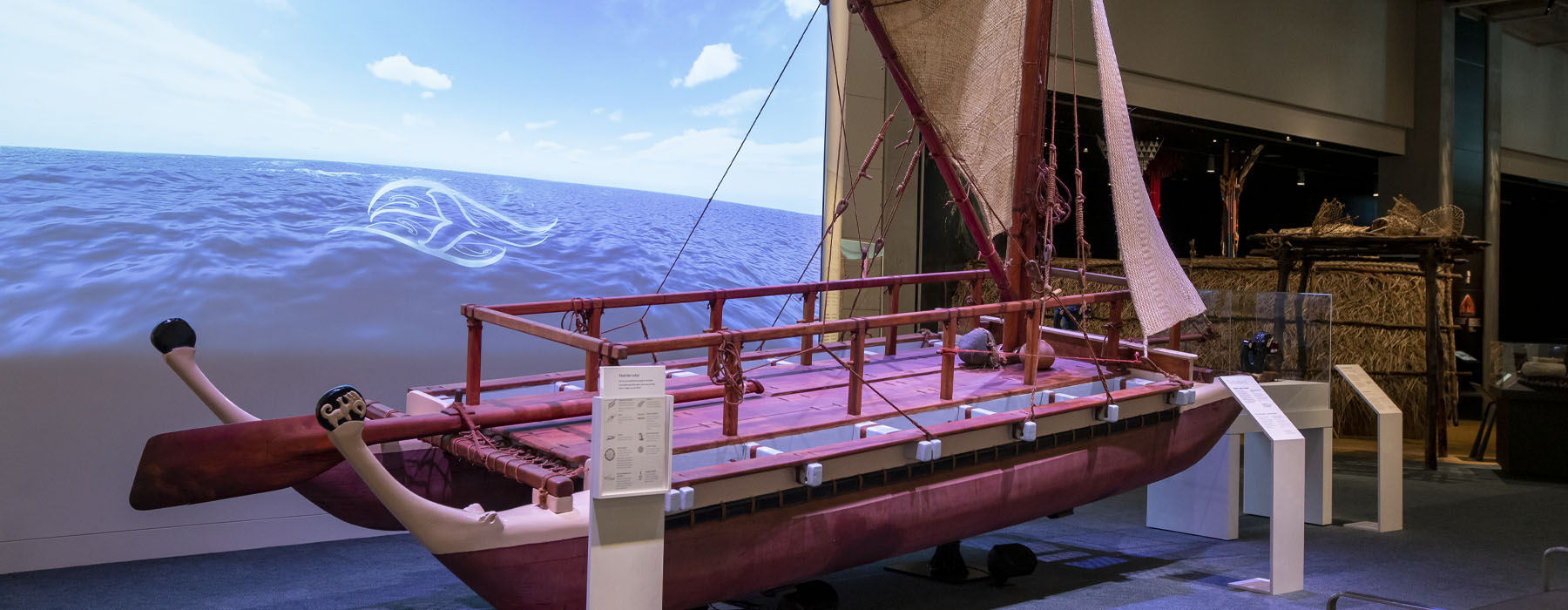 General view of an exhibition space with a large projection on the wall, a waka (Māori canoe) in the foreground, and further exhibits in the background