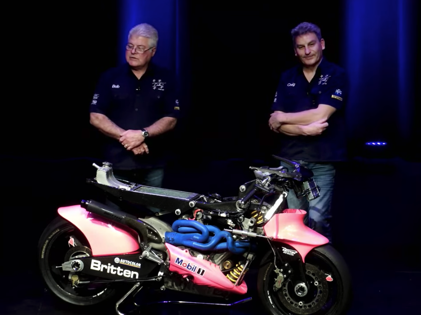 Still from the 2022 Britten Bike talk with Bob Brookland and Craig Roberts. Te Papa Two men standing on a stage with a bright blue and pink motorbike in front of them
