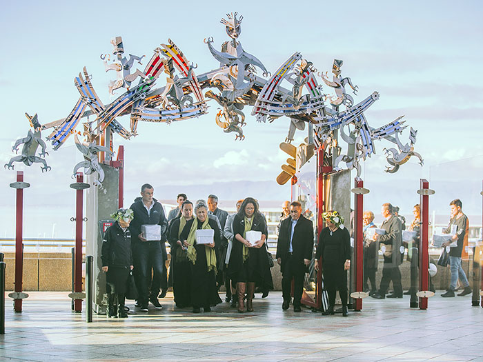 Group of people bring boxes containing the remains through the gates to the marae, with Wellington Harbour in the background