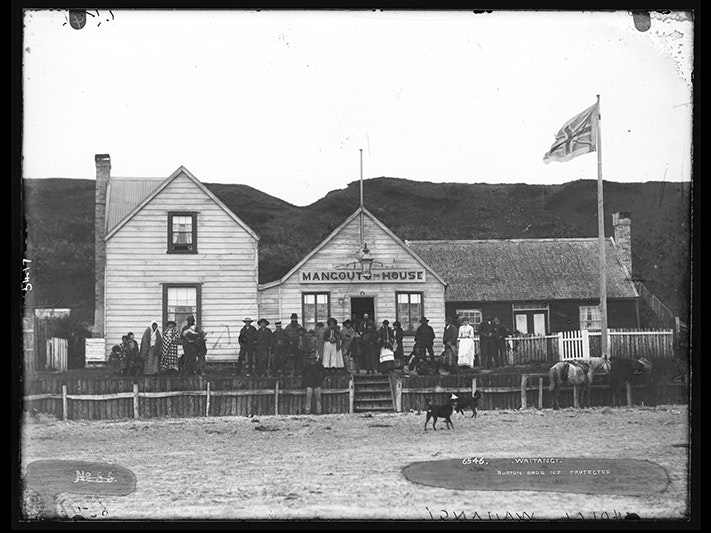 c.015417_waitangi.jpg A very old photo with people standing in front of a wooden building. There is a dog in the foreground.