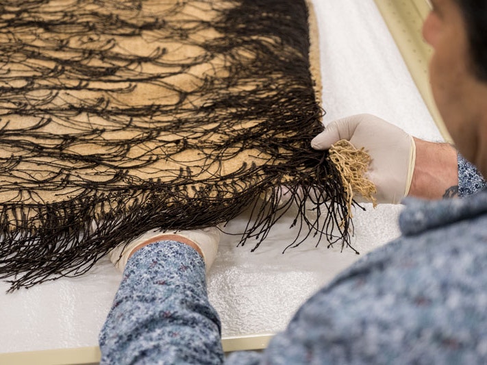Mark Sykes shows the hukahuka on a korowai, 2018. Photograph by Rachael Hockridge. Te Papa A person with gloved hands is holding the corner of a Māori cloak on a drawer.