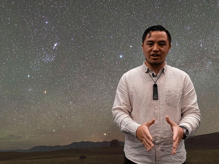 Still from How to find the Matariki star cluster, 2022. Photo by Daniel Crichton-Rouse. Te Papa Te Papa staff member Hauiti Gardiner talks to the camera. Behind him is a photograph of the night sky