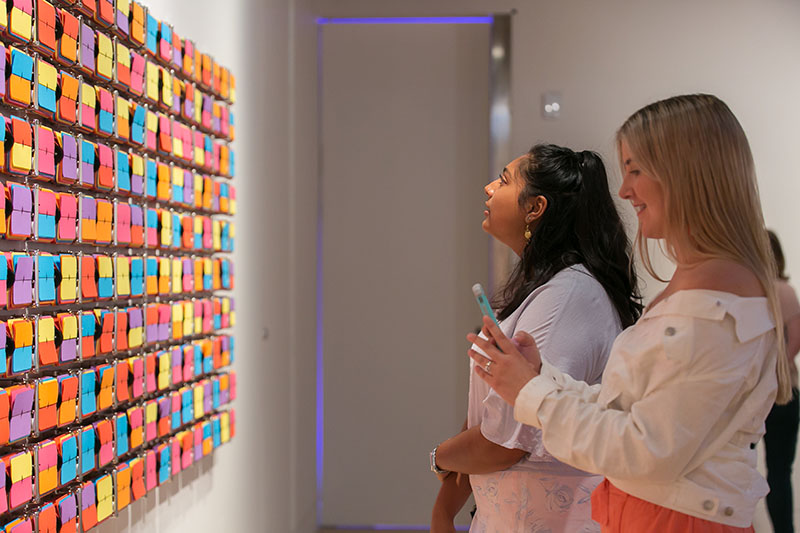 Two women stand in front of Rebecca Baumann’s Automated colour field (Variation 8)