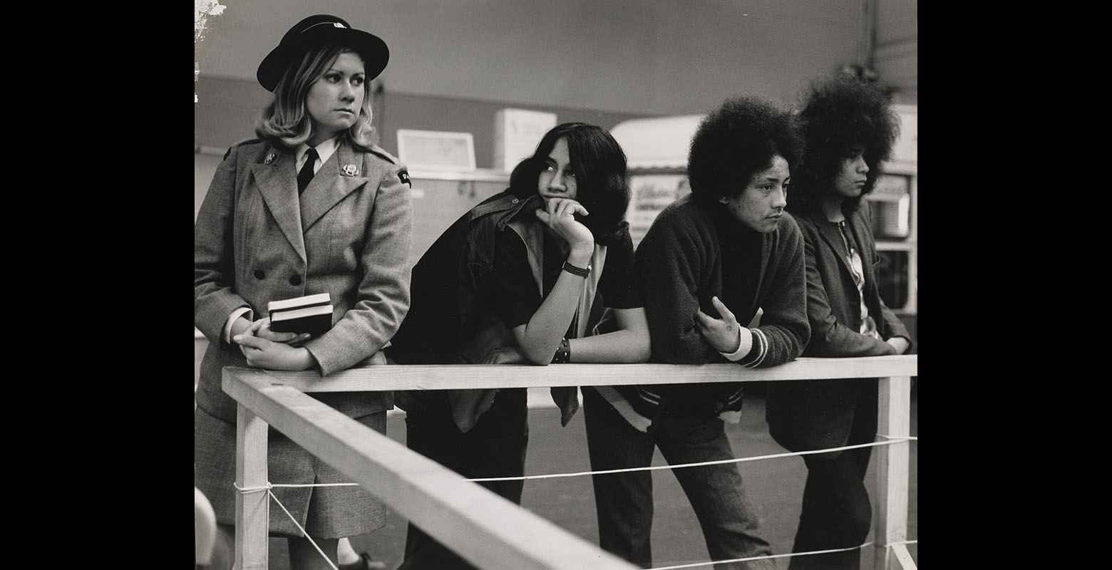 View of three young Māori men leaning on a wooden rail. On their left stands a young blonde woman in a St Johns uniform.