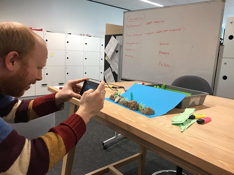 Donald photographs the model of the foreground image on a blue background. Photo by Martin Langdon. Te Papa Man takes photo of a model of underwater landscape, featuring rocks and small trees and a blue background