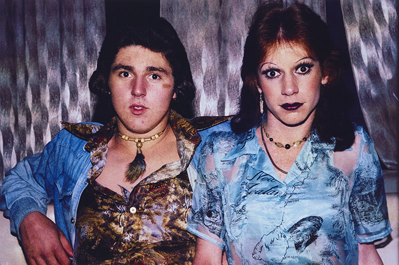 Two people seated look into the camera – one a man in an open shirt with a forest on it and denim jacket, the other a man in makeup wearing a blue shirt with a map of New Zealand on it