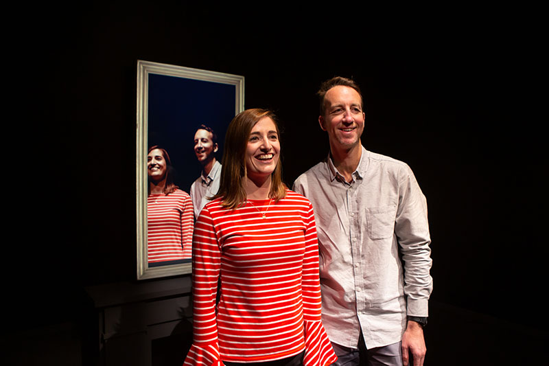 A man and a woman, smiling, look beyond the camera with their backs to a mirror. The mirror showing what we see, an impossible view of them looking in the same direction