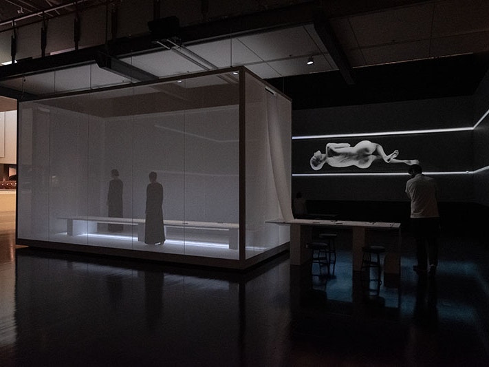 Installation view of Lemi Ponifasio, MAU: House of Night and Day, 2019. Photo by Rachael Hockridge. Te Papa A man walks around a space that contains a box with two women inside and a massive projection on the wall of a woman contorting her body