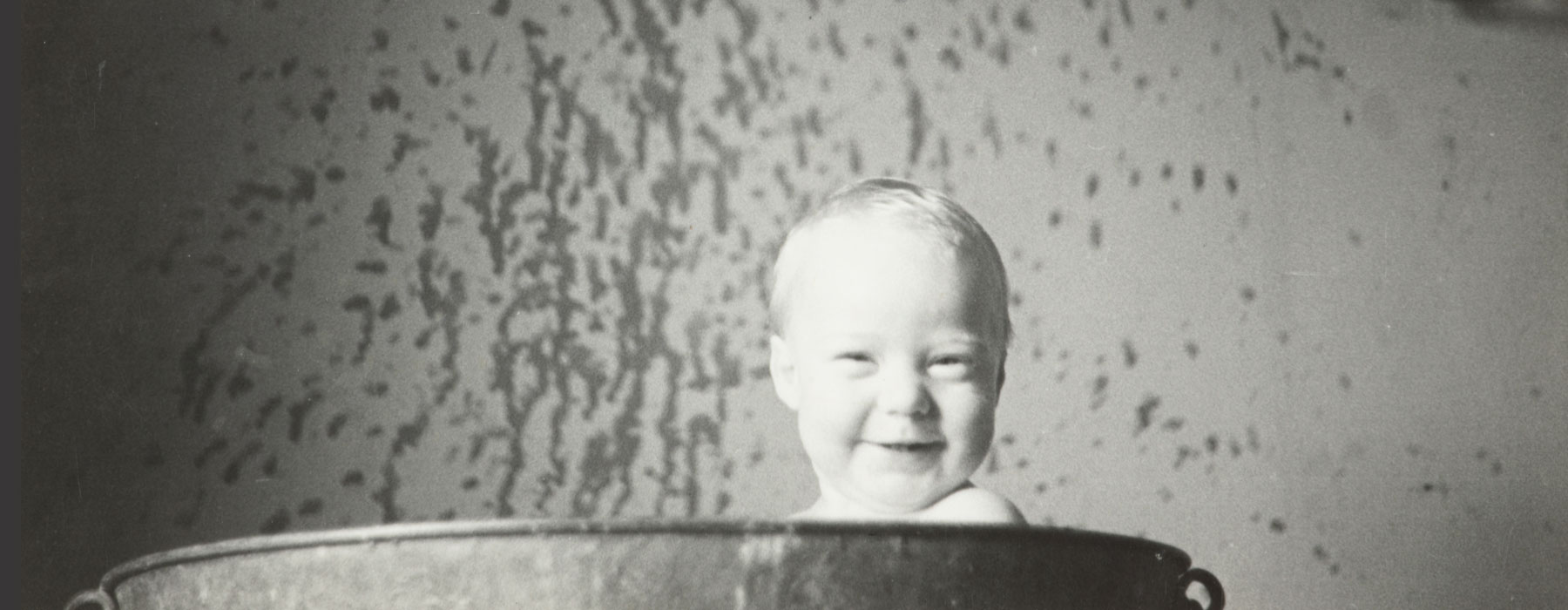 A baby grins, he's poking his head above a bathtub. There's water splashes on the walls.