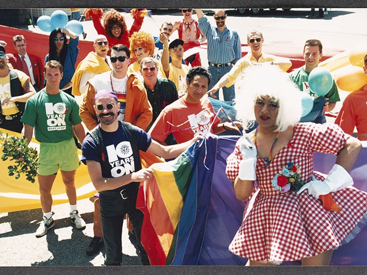 Devotion Festival, 1993, from Johnny Croskery’s photograph album 1980s–1990s, photographer unknown, New Zealand. Gift of Peter Kooiman, 2016. Te Papa (O.044266) Group of people in an assortment of of costumes stand in a parade holding the rainbow flag