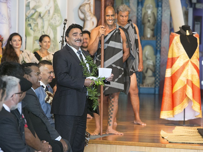 Te Papa Kaihautū Arapata Hakiwai speaks at the ceremony to return the ‘ahu ‘ula and mahiole, 2016. The cloak lies flat while a replica is draped on a mannequin. Te Papa Arapata addresses the room standing in front of a cloak draped on a mannequin