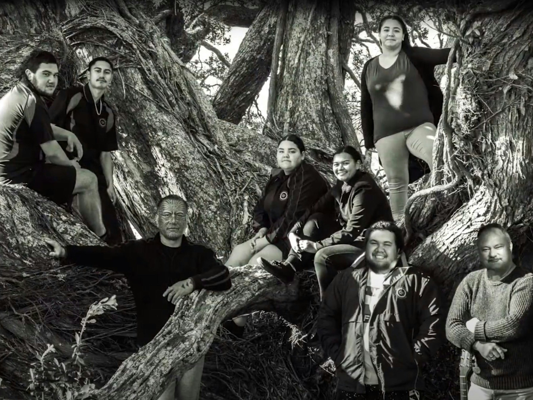 A still from the video for the song Mokohuruhuru. A group of people facing the camera sitting in and standing in front of a tree