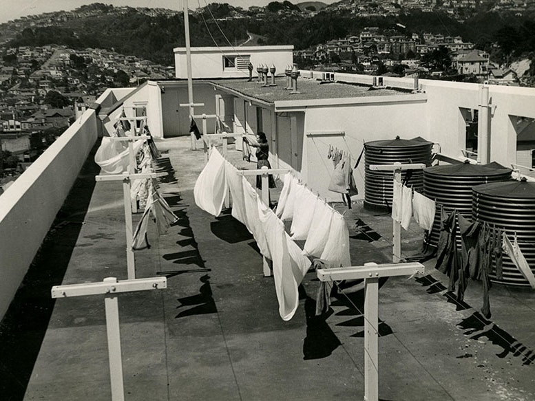 Dixon St flats, Wellington, about 1940, photographer unknown. CC BY 2.0. Archives New Zealand via Flickr (ABVF 7484 W4925 Box 1/ 1/17) Black and white photo of a woman hanging out the washing on the roof of an apartment complex