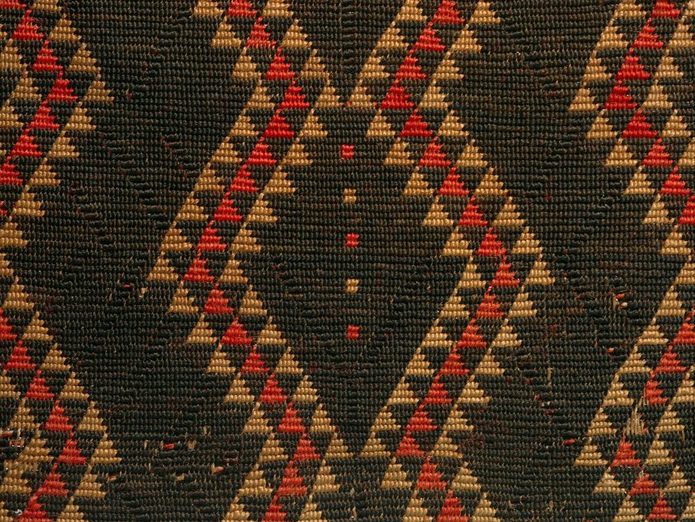 Close up of a cloak, showing the detail of its design. It is predominantly black with red and earth coloured triangles forming a wave across the cloak