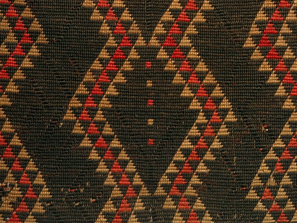 Detail of kaitaka paepaeroa with ngore (cloak with taaniko borders and vertical aho, weft rows), date unknown, maker unknown. Te Papa (ME015672) Close up of a cloak, showing the detail of its design. It is predominantly black with red and earth coloured triangles forming a wave across the cloak
