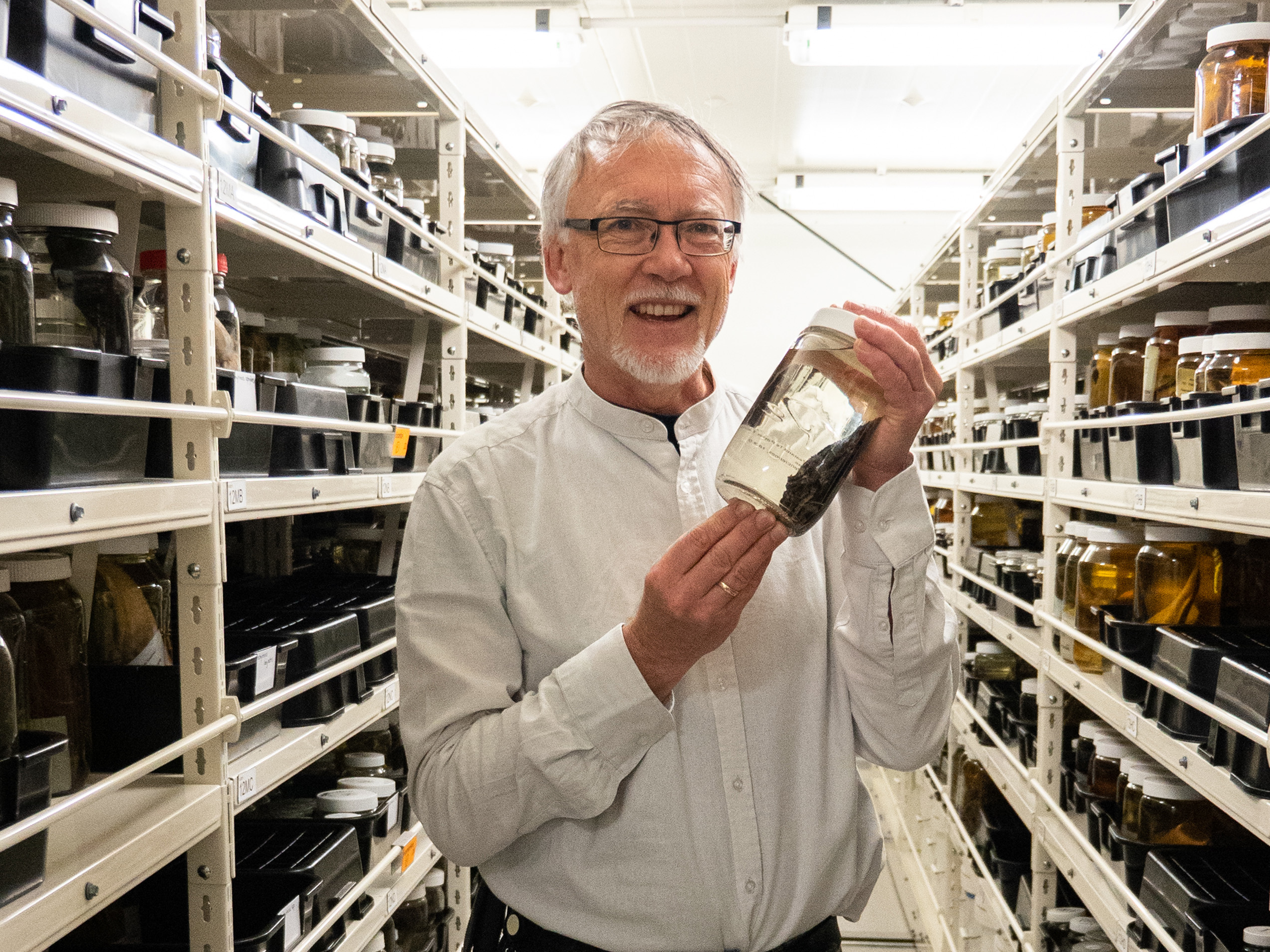 A man in a room of shelves holding a jar with a fish in it