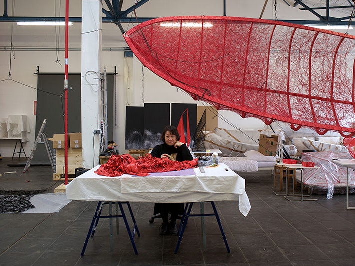 Chiharu Shiota, 2020. Photo by Sunhi Mang. © Copyright Agency, 2020 and the artist Portrait of Chiharu Shiota sitting at her desk in her studio. Above her hangs the red frame of a sailing boat frame with red wool being threaded across it