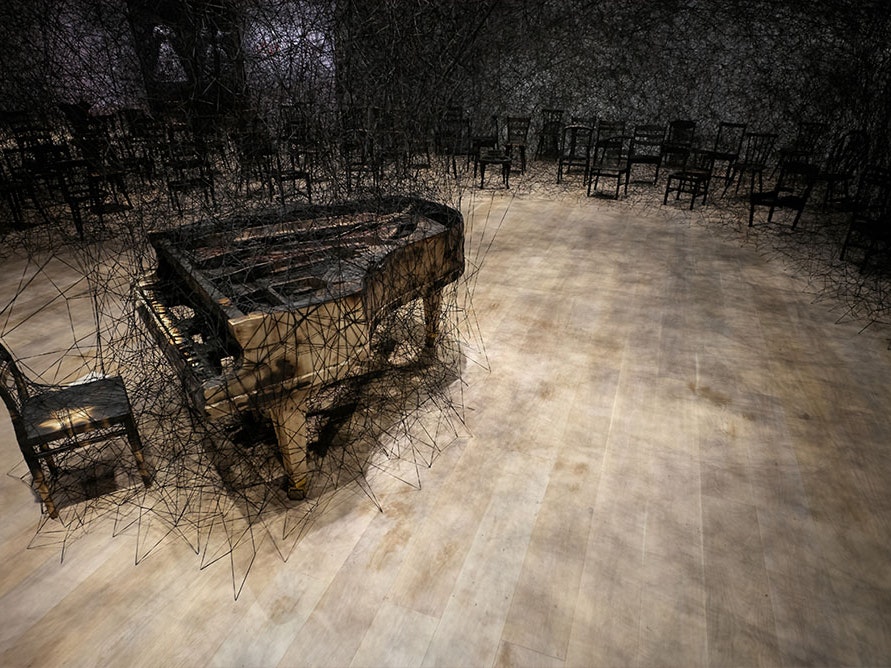 In Silence, 2019. Installation: burnt piano, burnt chair, Alcantara black thread. Installation view: Shiota Chiharu: The Soul Trembles, Mori Art Museum, Tokyo, 2019. Photo by Sunhi Mang. Courtesy of the Mori Art Museum, Tokyo. © Copyright Agency, 2020 and Burnt piano and chair stand in a room. Behind them are an array of chairs connected to each other and the stairs via black wool