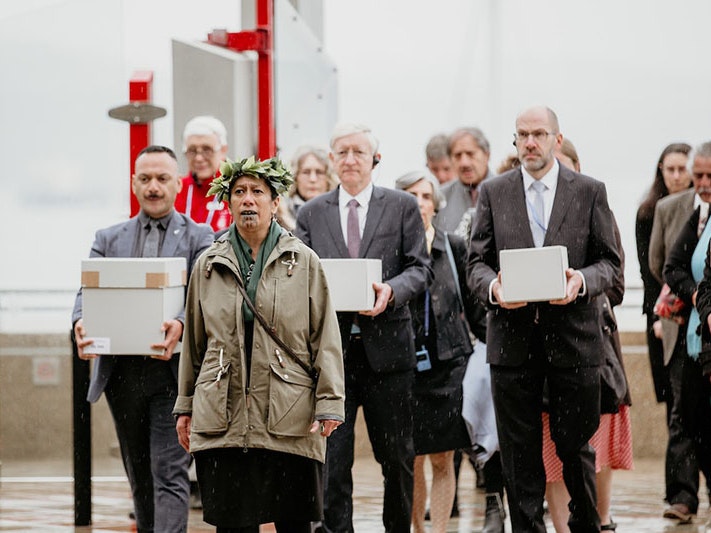 The delegation prepares to enter Te Papa’s marae. Te Papa’s Lisa Osborne gives the traditional karanga (call). Visible carrying three of the four toi moko are (left to right) Te Papa Repatriation Coordinator Te Arikirangi Mamaku, German Ambassador to New A group of people enter a building. Three of them carry boxes