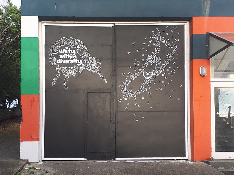 Mural on Daniell Street, Wellington, 3 April 2019. Photo by Katie Cooper. Te Papa (136502) A black sign on a garage that has a white kiwi and a map of New Zealand drawn on it with the words "Unity within diversity"