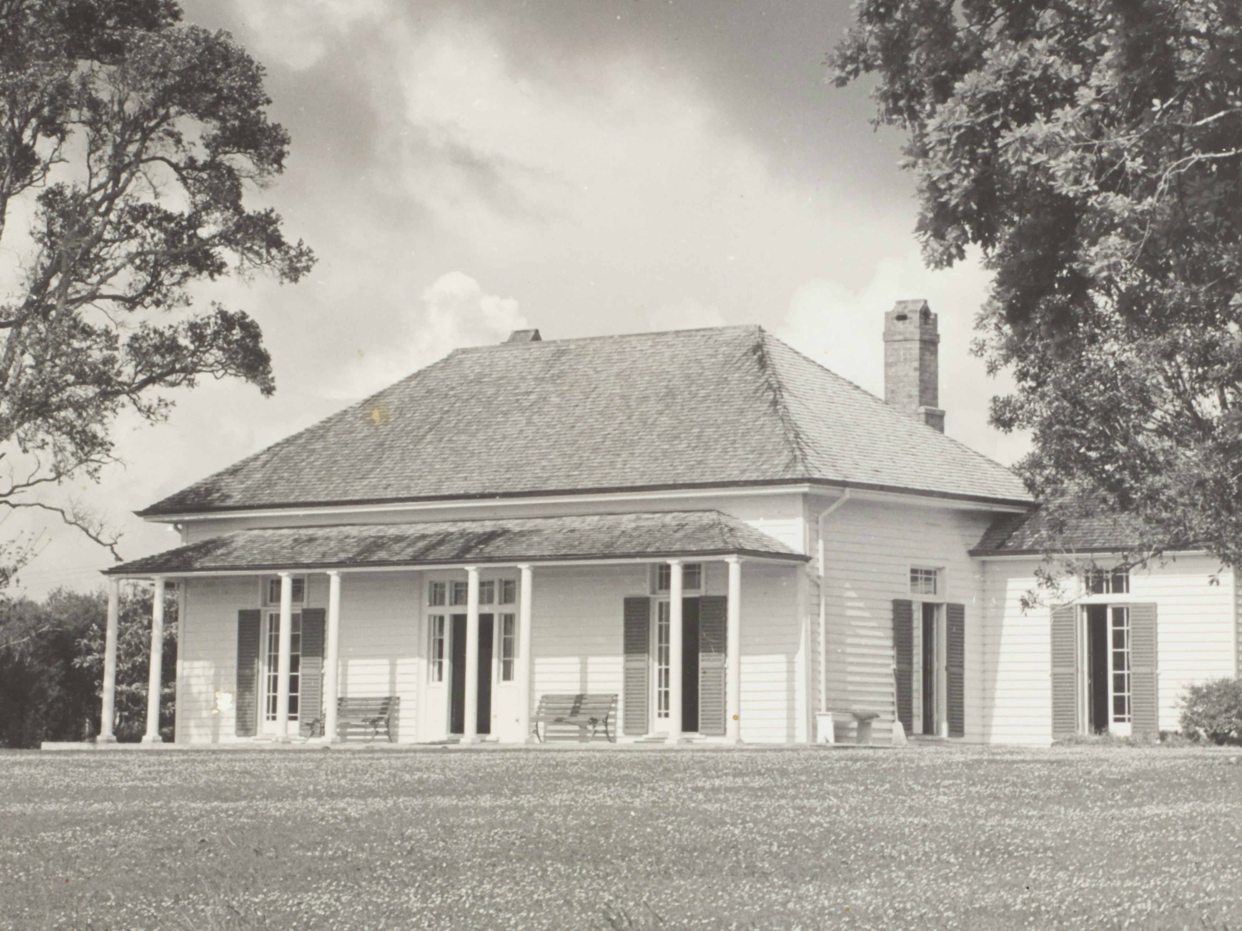 Treaty House, Waitangi, October 1956, Waitangi, by Eric Lee-Johnson. Purchased 1997 with New Zealand Lottery Grants Board funds. CC BY-NC-ND 4.0. Te Papa (O.010962) A one-storey wooden house in between two trees