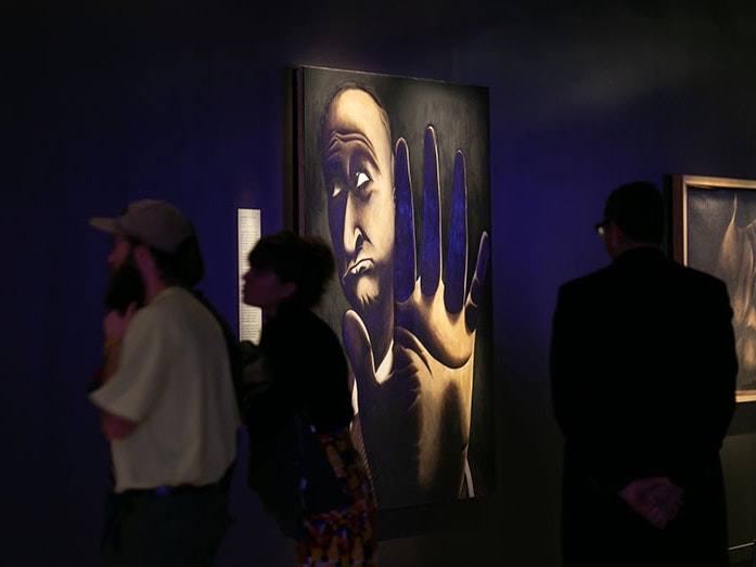Opening night of Tony Fomison: Lost in the Dark at Te Papa, 2018. Te Papa (114445) People look at a painting by Tony Fomison. The painting depicts a man holding his hand up to the camera as if to say No