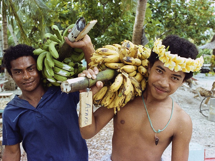 Glenn Jowitt, Men carrying bananas, Tokelau, 1981. Gift of Glenn Jowitt Estate, 2015. Te Papa (E.007344/18) Two men holding bunches of bananas balanced on their shoulders pose for a photo