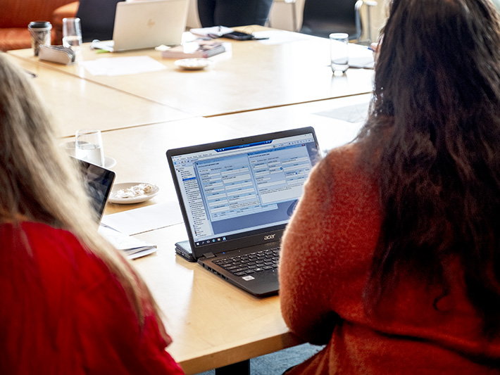 People gathered around a table with computers, there is a woman in the foreground looking at a screen.