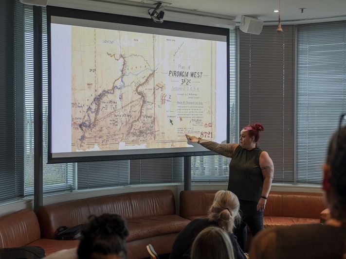 A woman is standing in front of a large screen and pointing at a place on a map in front of an audience.