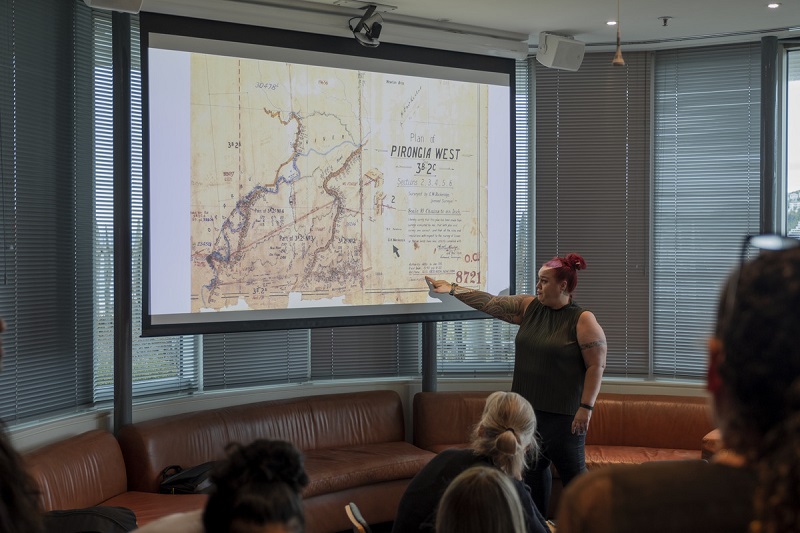 A woman is standing in front of a large screen and pointing at a place on a map in front of an audience.