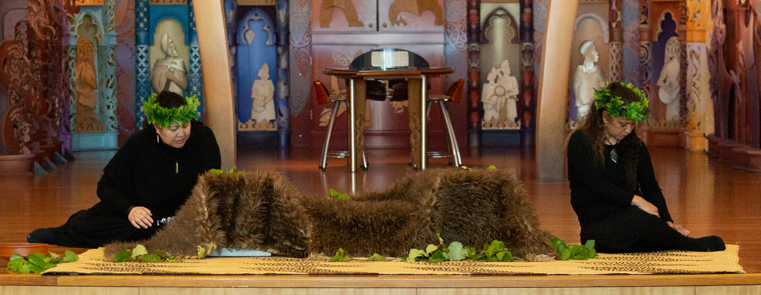 Two women in black with green-leaved wreaths on their heads sit on a stage looking at boxes covered in feather cloaks