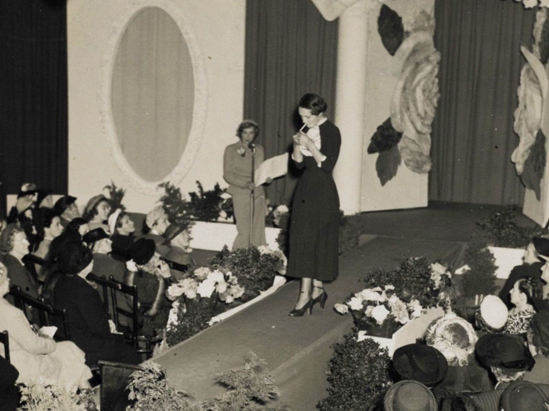Crop of a photograph from The Romance of Wool, a New Zealand Wool Board Mannequin Parade, November 1949. Te Papa (CA000911/001/0002/0009) A black and white photo of a woman lighting up a cigarette on the catwalk