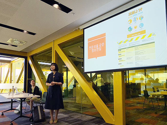 A woman sitting at a desk with another woman standing in front of a large screen showing Covid-19 signs in the Korean language.