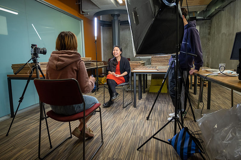 A woman sits on a chair in the middle of a room, being interviewed by another woman sitting opposite her. A large light shines on the subject