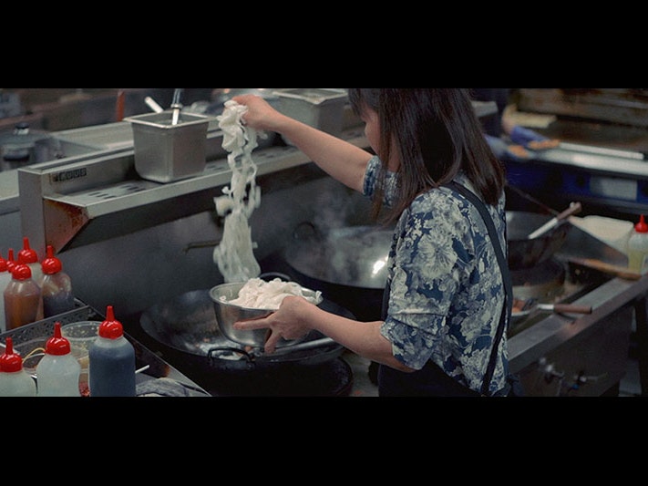 Still from the video, of Tee cooking at Little Penang, 2021. Photo by Arthur Hon Sheng. Te Papa A woman is seen from behind putting noodles into a wok in a busy kitchen