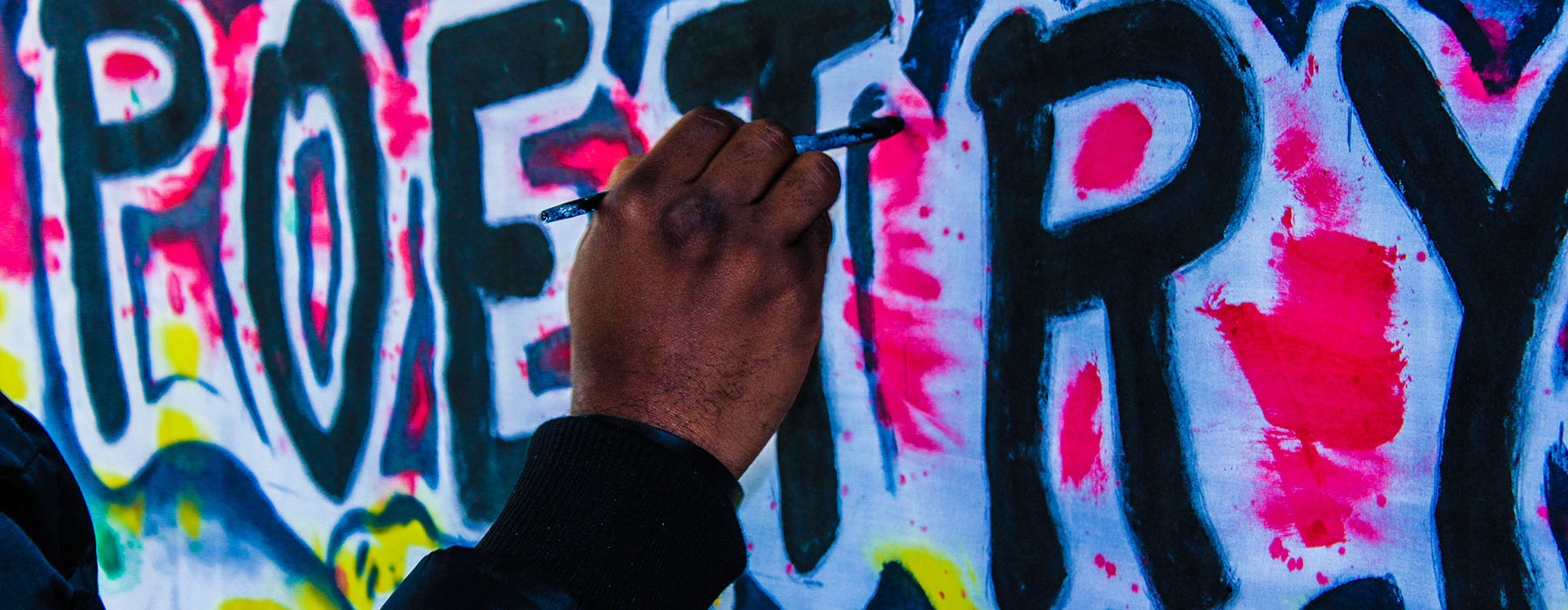 Text on wall that says “Poetry” being painted by a hand