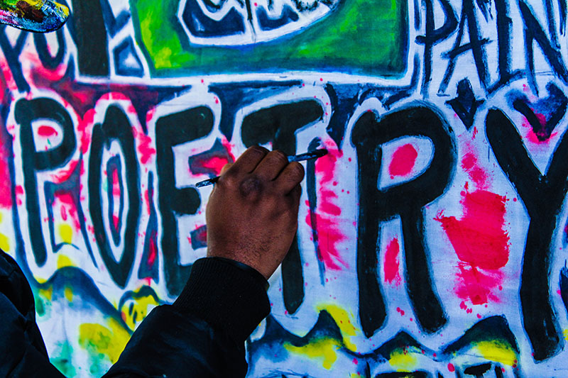 Text on wall that says “Poetry” being painted by a hand