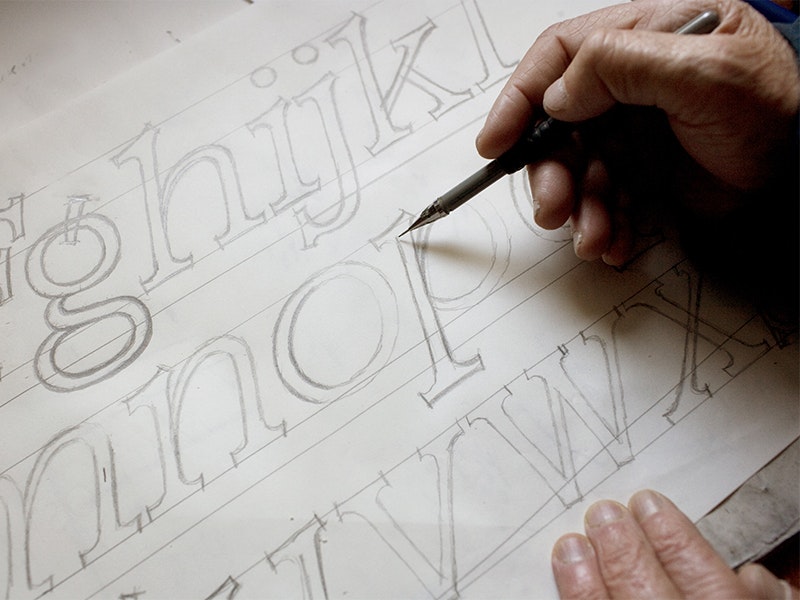 Detail of Joseph Churchward sketching at his desk, 2008. Photograph by "Norm Heke". Te Papa (65032) A close-up of a hand drawing caligraphy and another hand holding the piece of paper still.