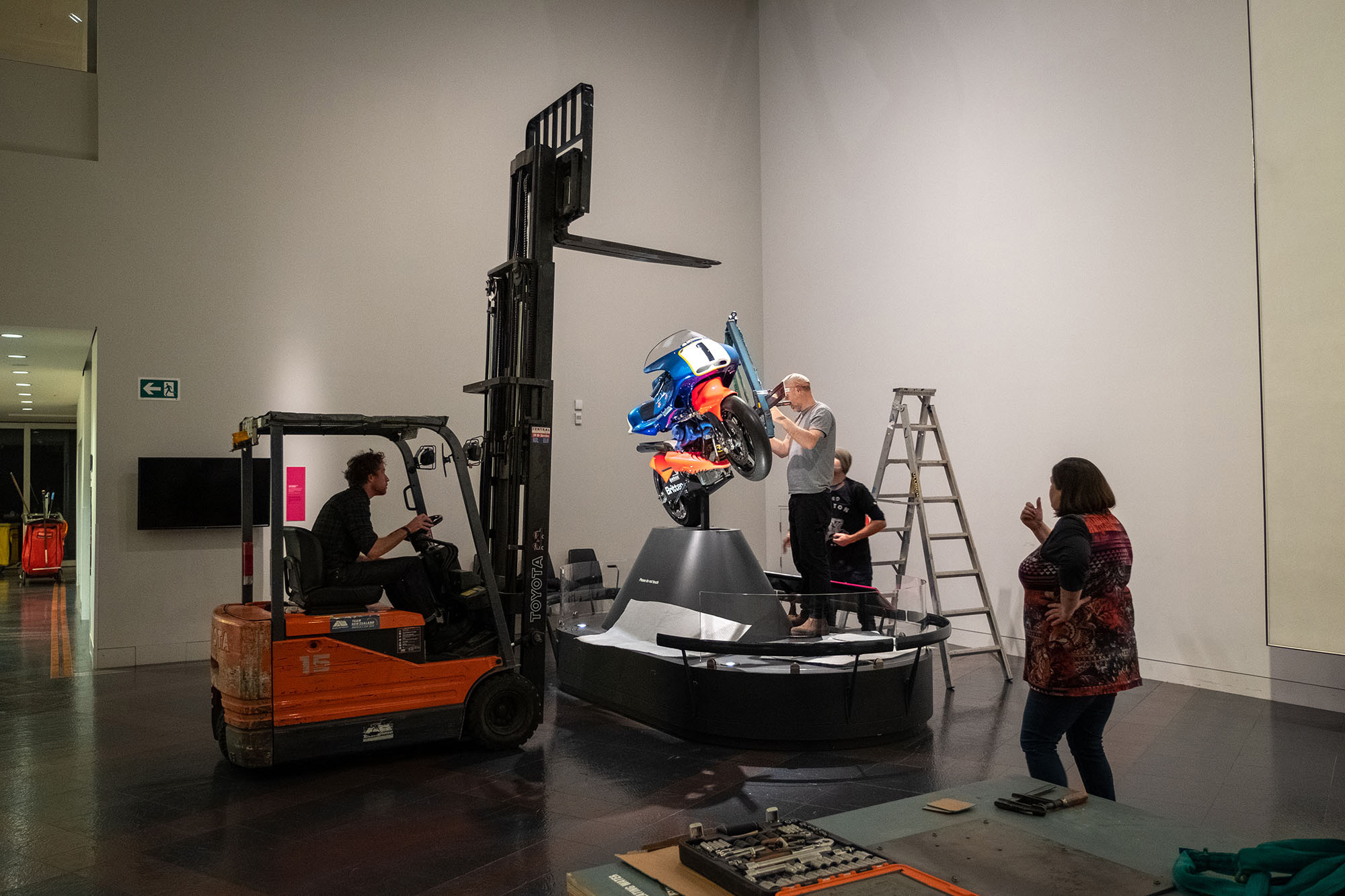 A man driving a forklift towards the Britten Bike display with people preparing for the bike to be lifted