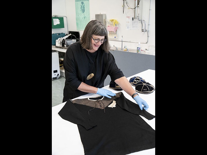 Claire Regnault with 1905 'Originals' All Black jersey, 2022. Photo by Maarten Holl. Te Papa (198903) Claire holds up for the camera a rugby jersey that is sitting on a table