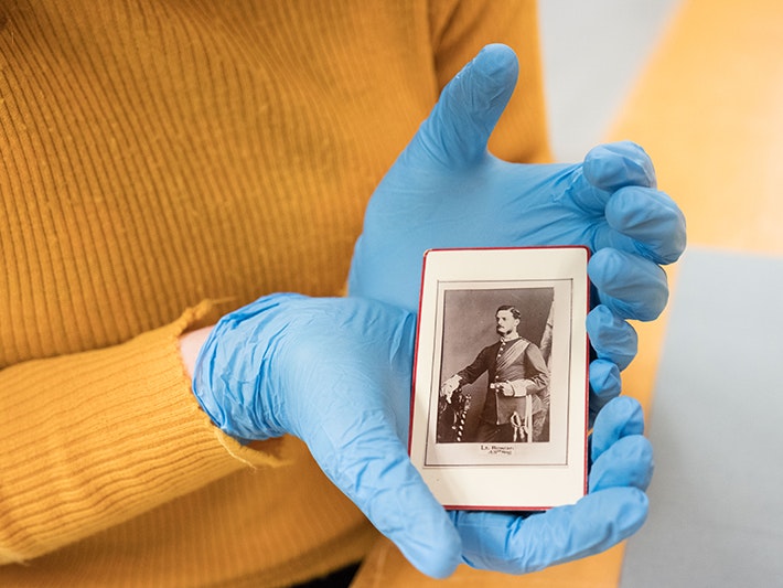 Caitlin holding a photograph of Frederick Rowan, 2018. Photo by Rachael Hockridge. Te Papa A pair of blue-gloved hands holding a small portrait photo in front of a yellow jersey.