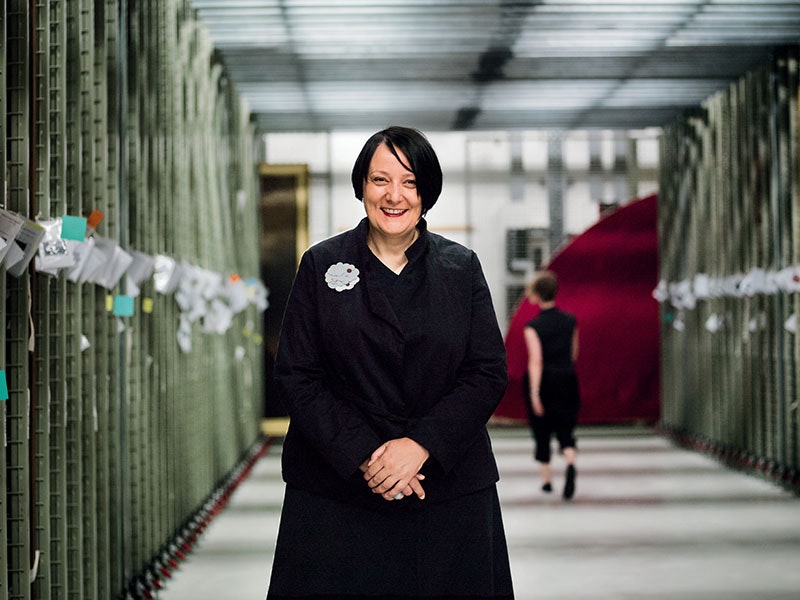 Head of Art Charlotte Davy in the art store room, 2017. Photo by Jane Ussher. Te Papa Charlotte poses in front of the racks that house paintings. A woman walks past behind her