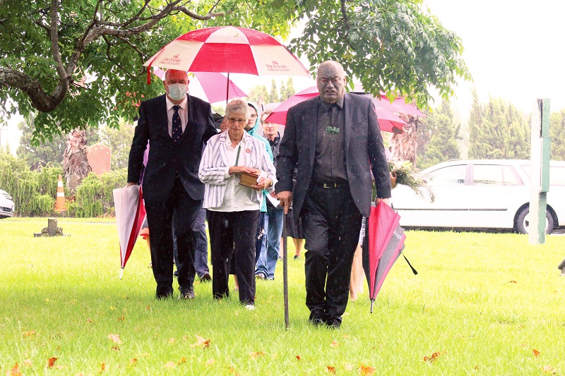 Some people carrying umbrellas and a woman carrying a wooden box are walking in a group towards the camera