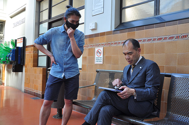 Two men talking. One is seated looking at something on an iPad. The other is standing, also looking at what is on the iPad.