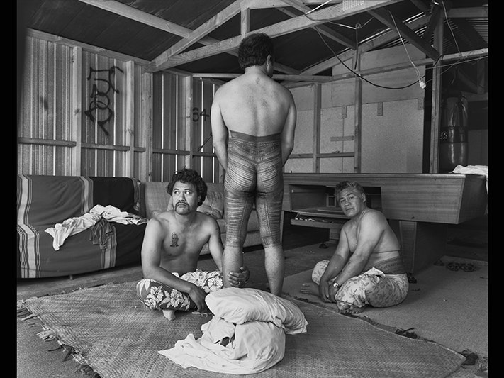 Mark Adams, Velvet Crescent, Otara, South Auckland, Fatu Feu‘u Tufuga tatatau: Su‘a Sulu‘ape Paulo II, 19 November 1991, print. Image courtesy of the artist. Three men with tattoos in a room. Two of the men are sitting on the floor, one is standing with his back to the camera