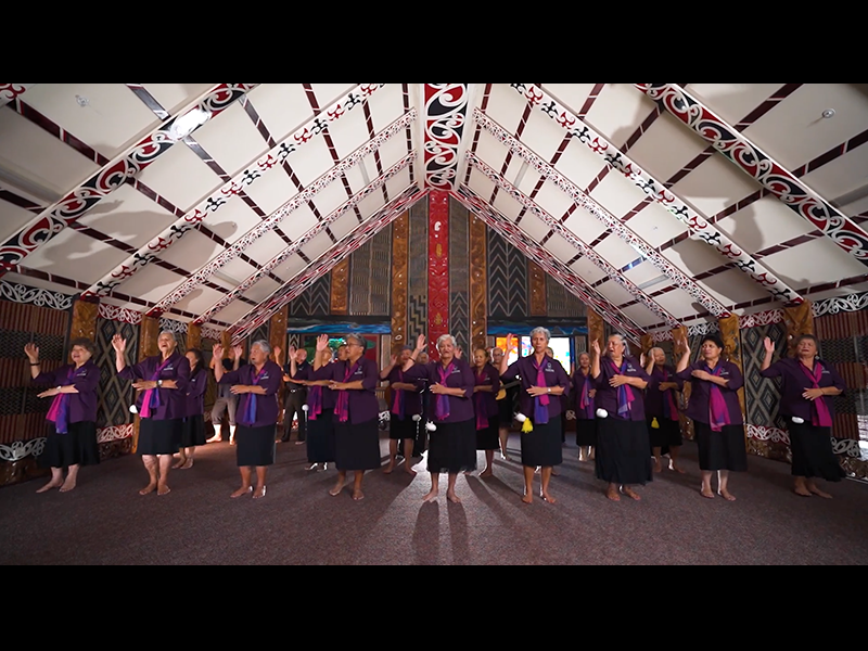 Video still from Taikura Kapa Haka 2022. Te Papa People in a group wearing the same clothing with pink and purple scarves facing the camera and singing with hand actions